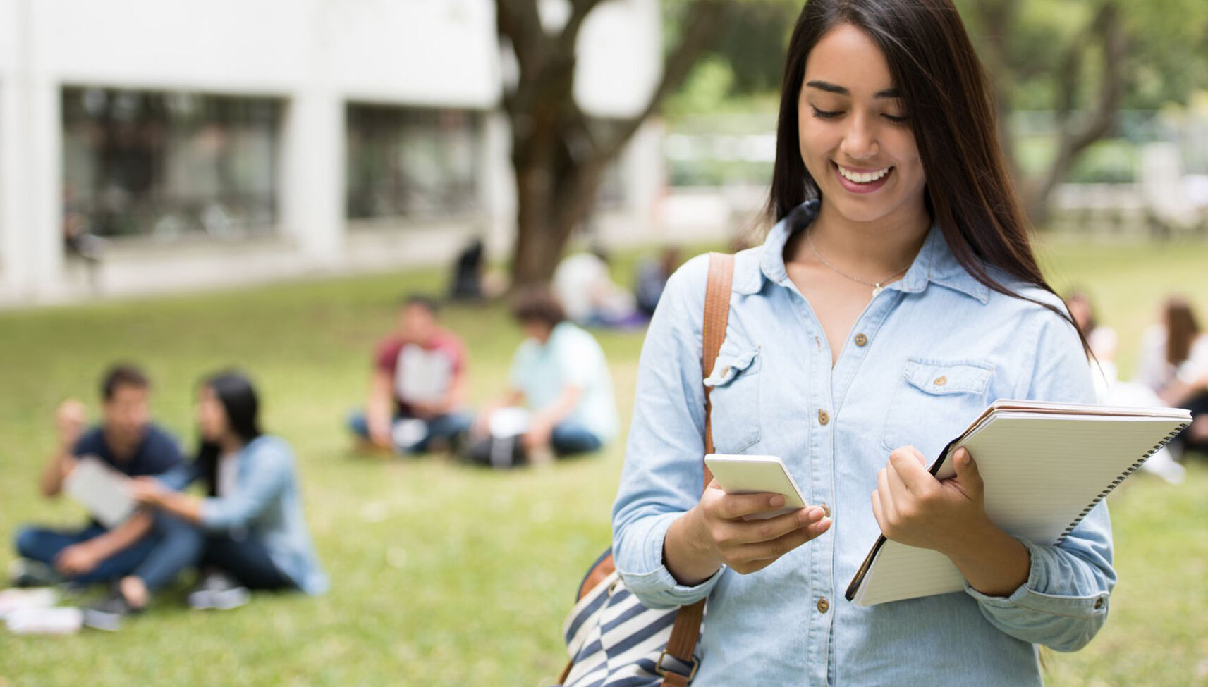 A female student walking outdoors holding her books and looking at her phone